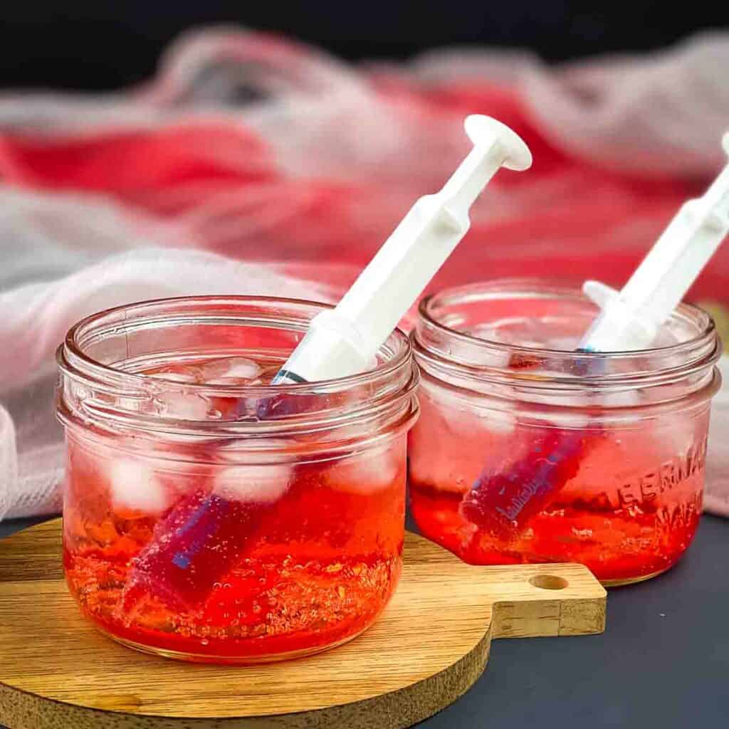 A small jar filled with a bloody shirley temple, with a plastic syringe in it.