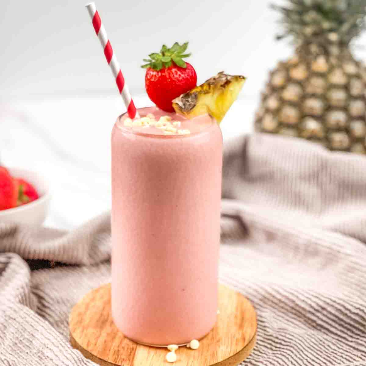 A tall glass of Bahama Mama tropical smoothie garnished with a whole strawberry, pineapple wedge, white chocolate chips, and a red and white striped straw.