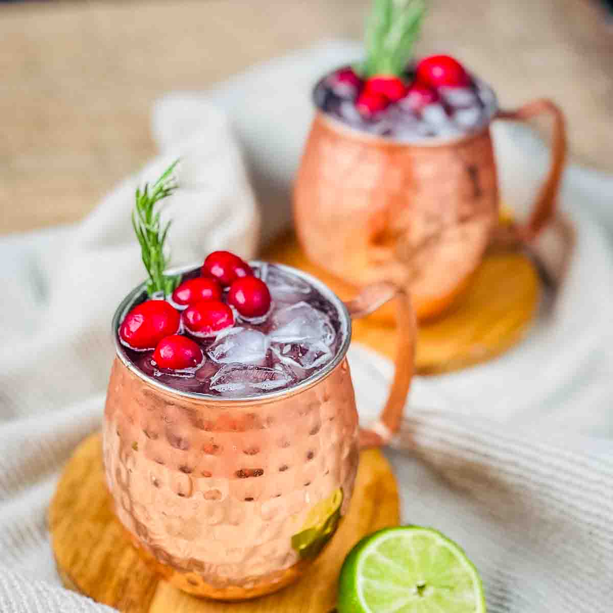 Two copper mugs of a winter mule, garnished with cranberries and a sprig of rosemary.