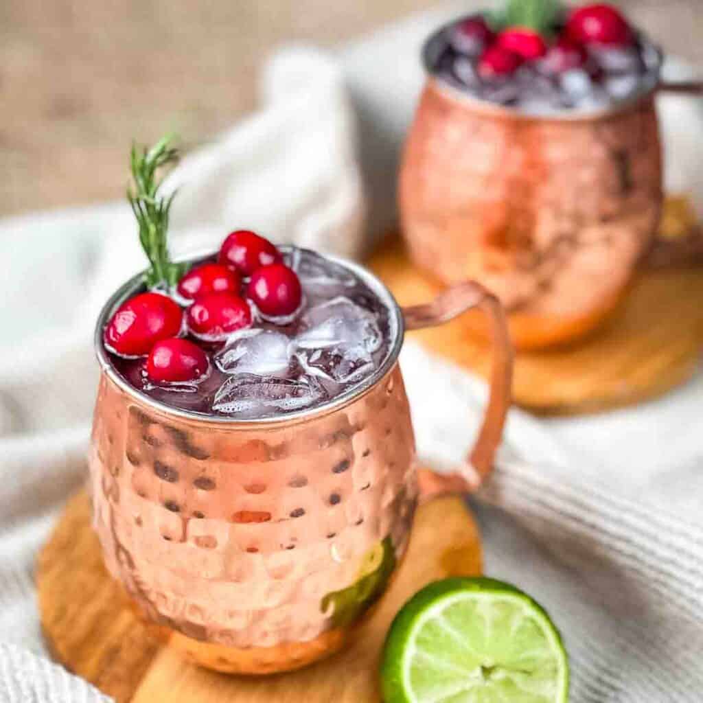 Two copper mugs of a cranberry Moscow mule garnished with fresh rosemary and cranberries.