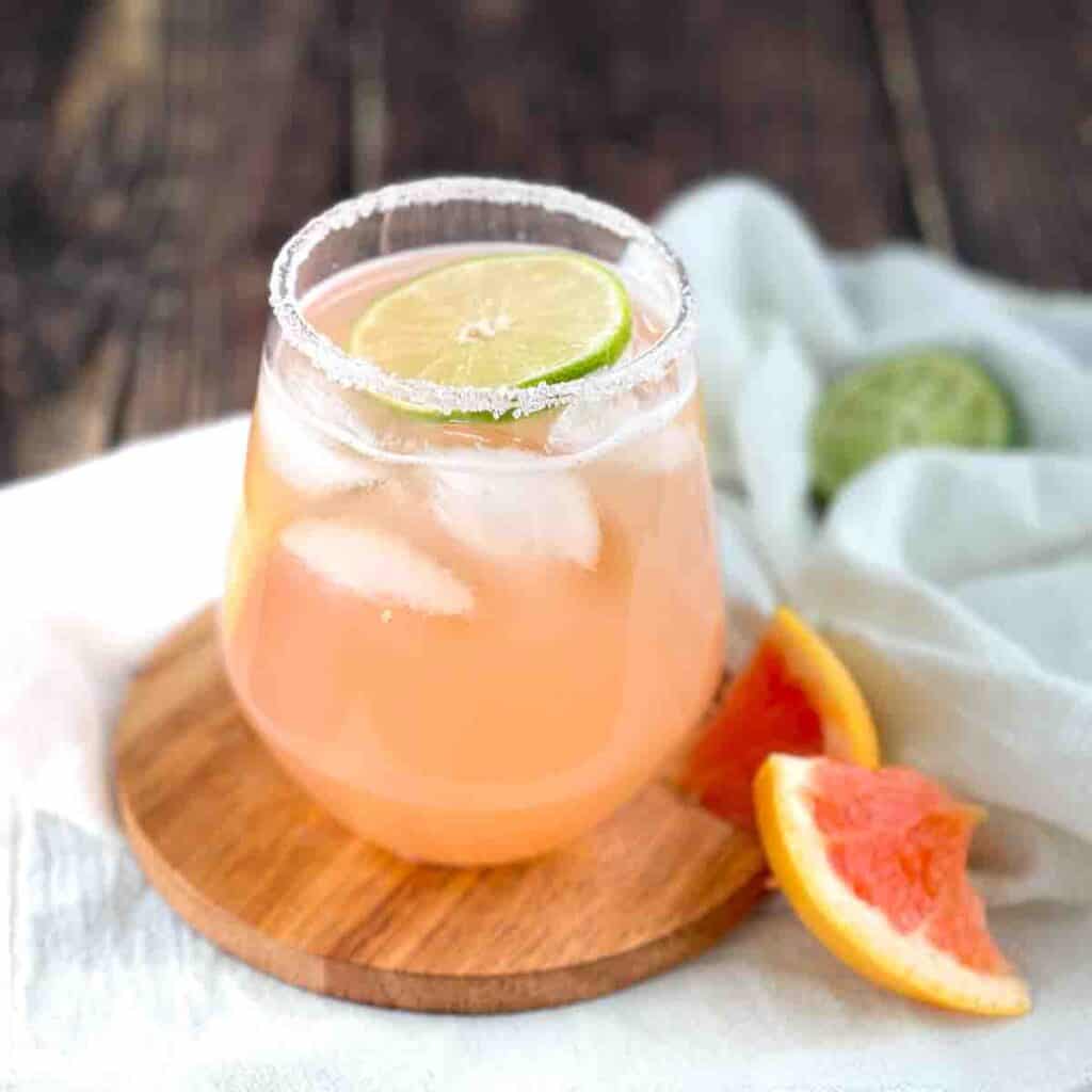 A grapefruit paloma in a stemless wine glass, garnished with a lime wheel and grapefruit wedge.