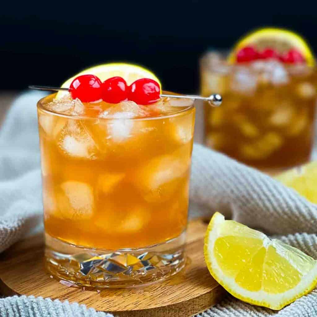 Two rocks glasses with a rum sour cocktail, garnished with maraschino cherries and lemon wedges.