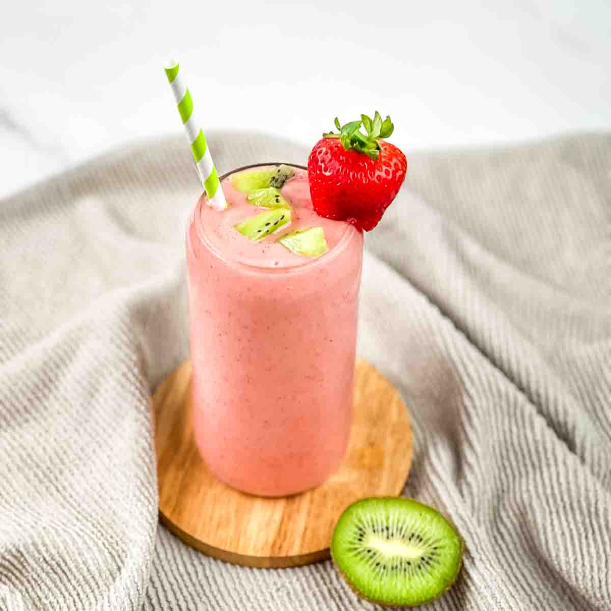 A tall glass filled with copycat tropical smoothie cafe kiwi quencher, garnished with fresh kiwi and strawberry, with a green and white striped straw.