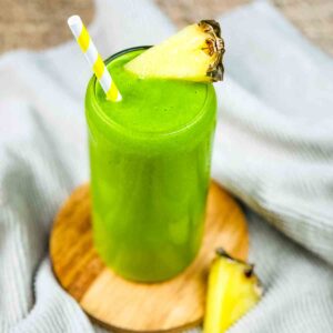 A vibrant green smoothie served in a tall glass, garnished with a fresh pineapple wedge with a yellow and white striped straw.