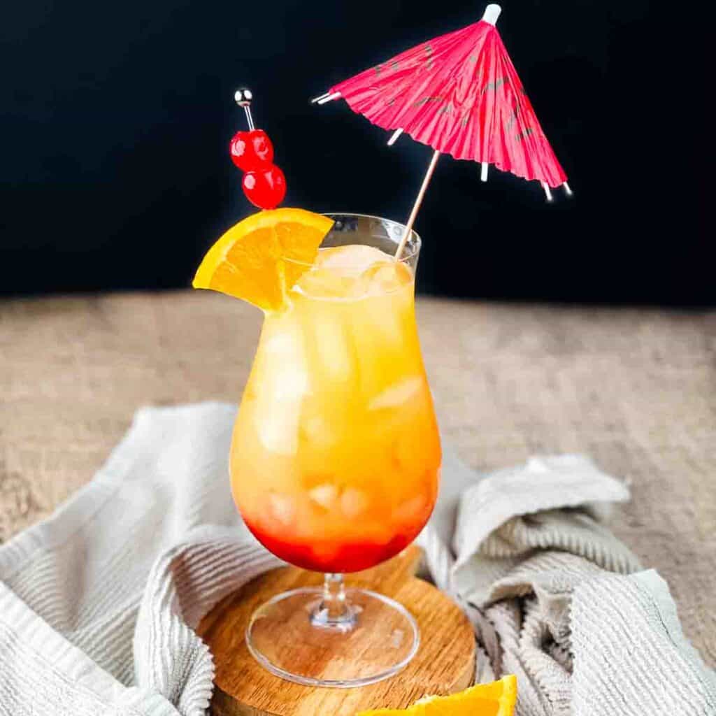 A hurricane glass filled with a sunset cocktail and garnished with an orange wedge, cherries, and an umbrella.