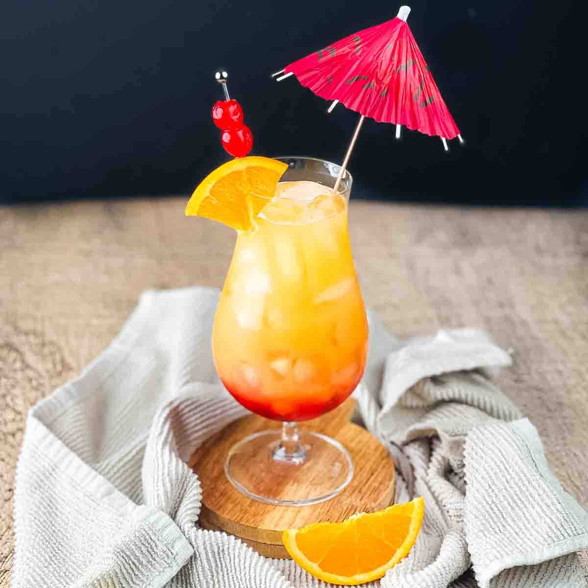 The sunset cocktail in a hurricane glass garnished with an orange wedge, cherries, and an umbrella.