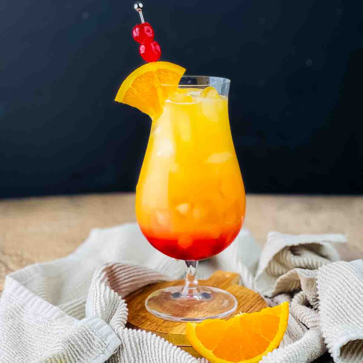 A sunset cocktail in a hurricane glass, showing off the sunset gradient of colors, garnished with an orange slice.