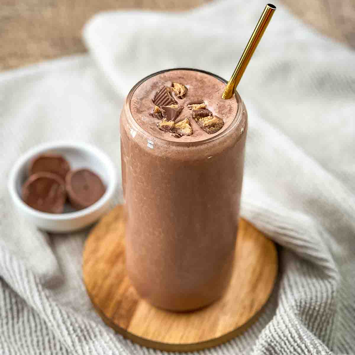 A peanut butter cup smoothie in a tall glass with a gold straw, garnished with crumbled peanut butter cups.