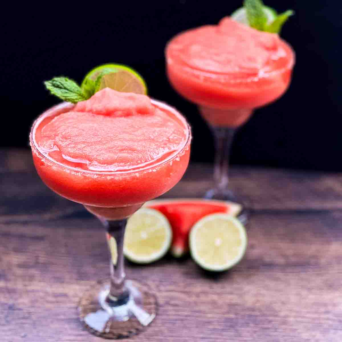 Two tall glasses of watermelon margaritas garnished with fresh mint and lime wedges.
