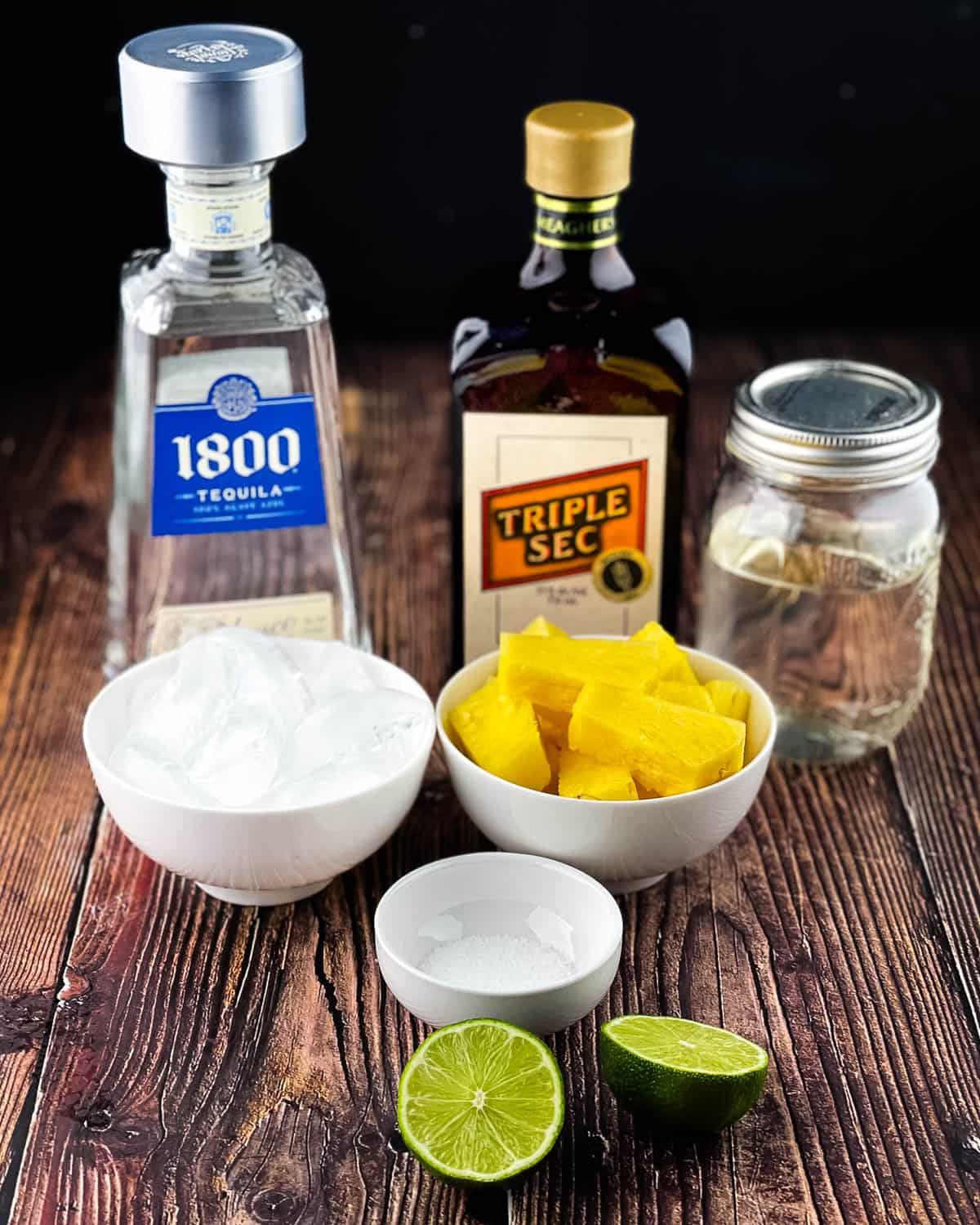 Ingredients for frozen pineapple margarita, including frozen pineapple, tequila, triple sec, lime, simple syrup, and ice arranged on a countertop.