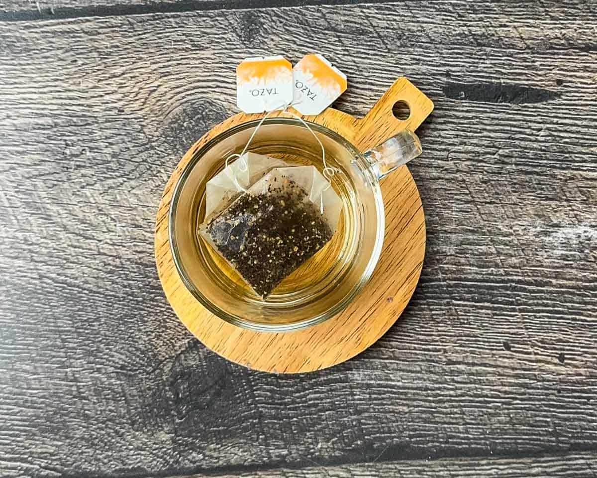 Two Tazo Chai tea bags steeping in a small amount of hot water in a clear mug to create a concentrated tea base.