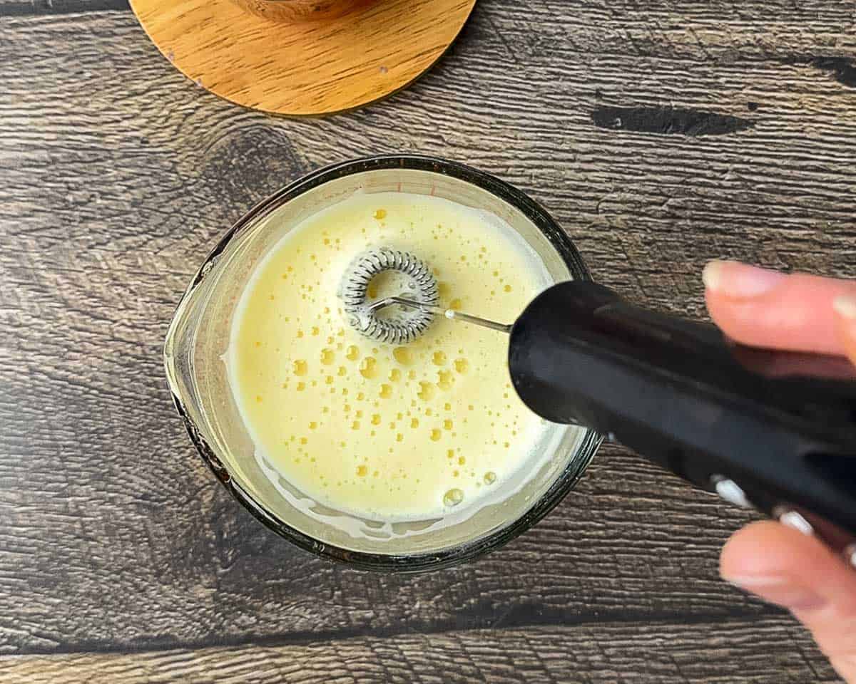 Handheld milk frother creating a thick, airy foam in a cup of warm eggnog flavored with vanilla and nutmeg.