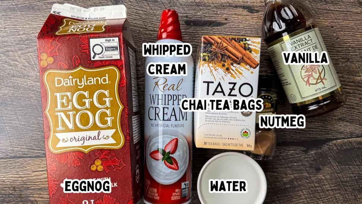A top-down shot of ingredients for an eggnog chai latte on a wooden table, including a carton of Dairyland Egg Nog, a can of real whipped cream, a box of Tazo Chai tea bags, a bottle of vanilla extract, a small jar of nutmeg, and a white bowl of water. Each item is clearly labeled with white text overlays.