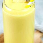 A tall glass filled with a creamy mango magic smoothie garnished with fresh pineapple and a yellow and white striped straw.
